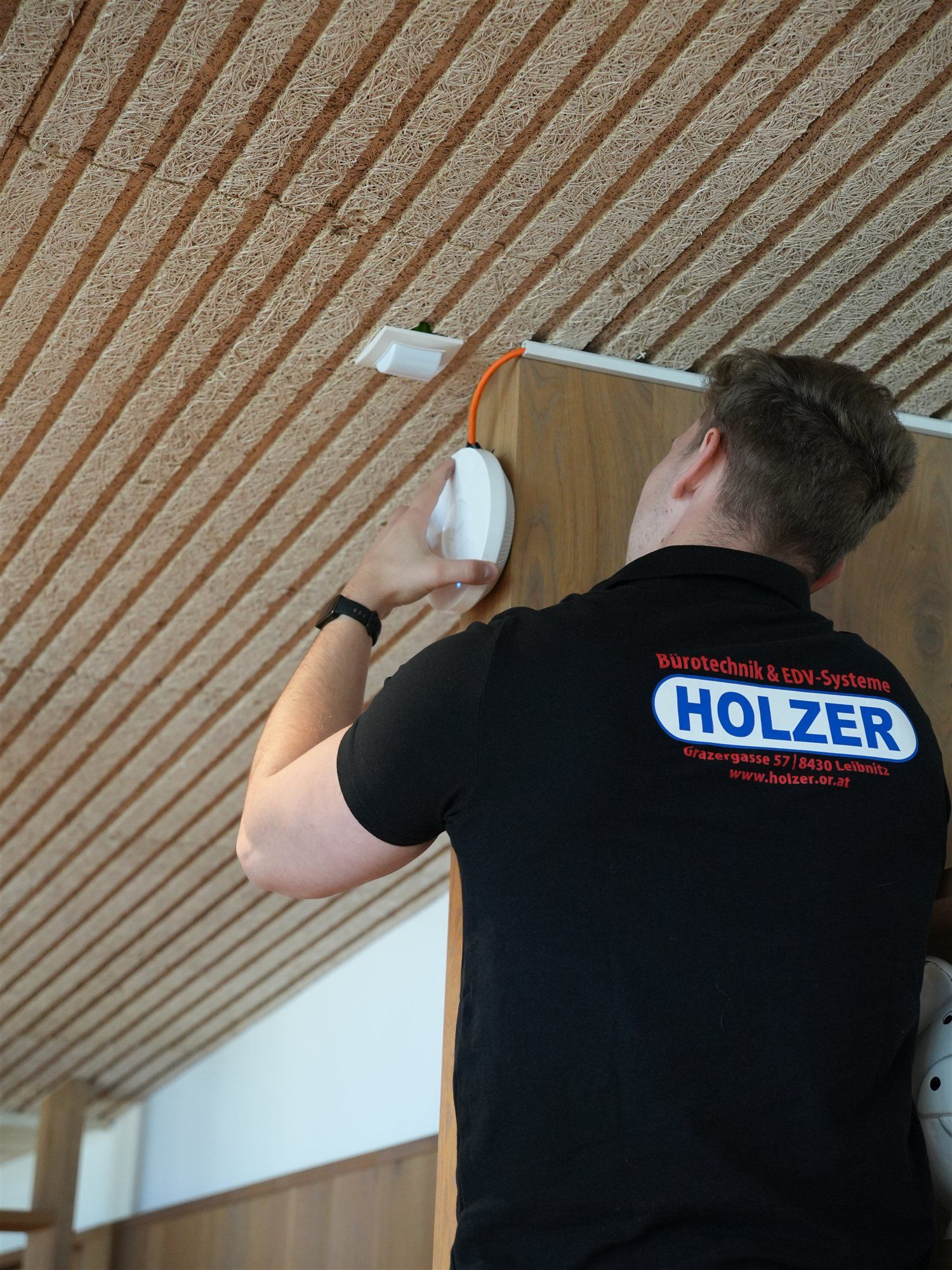 Holzer-Techniker montiert einen WLAN-Access-Point knapp unter der Decke, von hinten mit dem Holzer-Logo auf dem Shirt